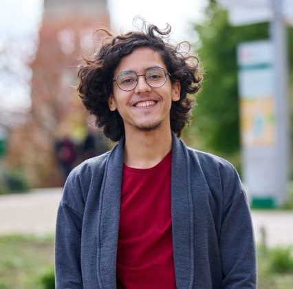 A smiling student with dark hair curly hair and glasses. They are wearing a long grey jacket, red t shirt and tan trousers. Behind them is a blurred background of a path through a park.